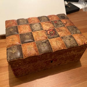 K&ouml;nnte beinhalten: Eine kleine, rechteckige Schachtel mit einem Schachbrettmuster auf dem Deckel. Die Schachtel ist aus Holz und hat eine rotbraune Farbe. Der Deckel ist mit einem goldfarbenen Design verziert.