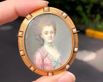 May include: A gold-toned oval brooch featuring a miniature portrait of a woman in a pink dress with a blue bow. The brooch is framed with small pearls and a decorative border, held by a hand.