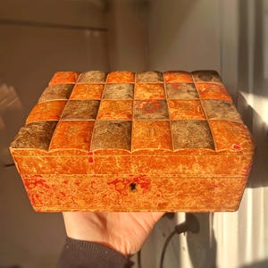 K&ouml;nnte beinhalten: Eine rechteckige Holzkiste im Vintage-Stil mit einem Schachbrettmuster auf dem Deckel. Die Kiste ist aus hellem braunem Holz mit einer r&ouml;tlich-braunen Beize. Der Deckel hat ein kleines Schl&uuml;sselloch in der Mitte.