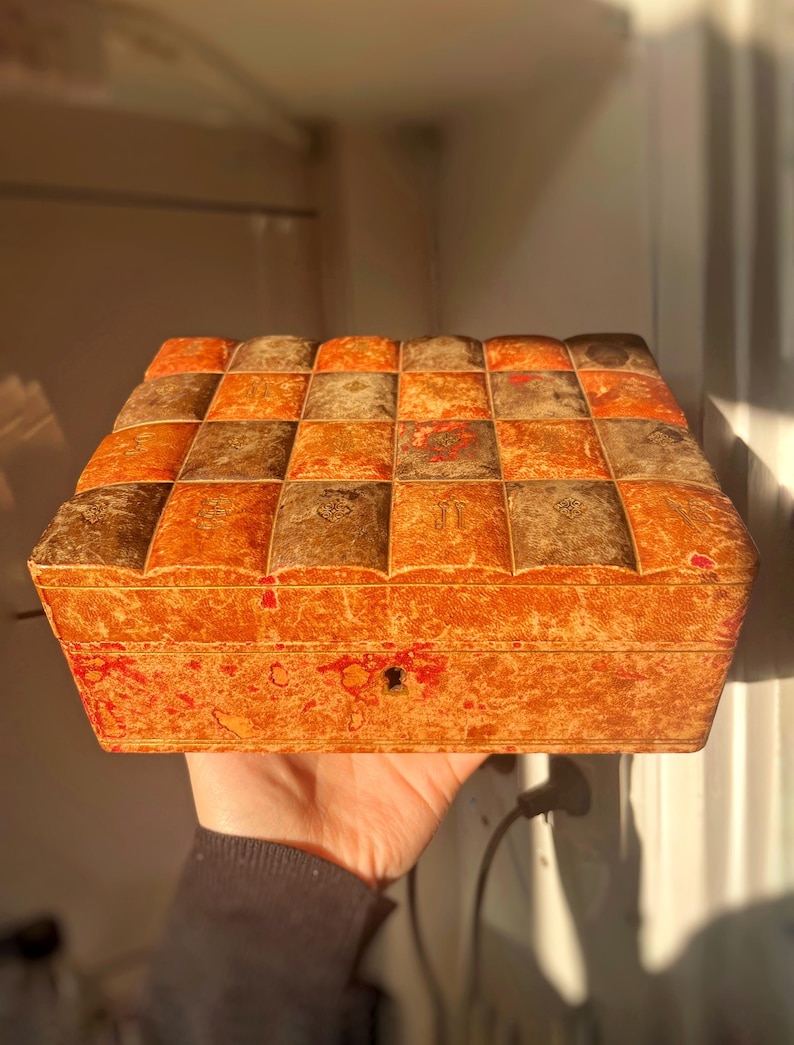 K&ouml;nnte beinhalten: Eine rechteckige Holzkiste im Vintage-Stil mit einem Schachbrettmuster auf dem Deckel. Die Kiste ist aus hellem braunem Holz mit einer r&ouml;tlich-braunen Beize. Der Deckel hat ein kleines Schl&uuml;sselloch in der Mitte.