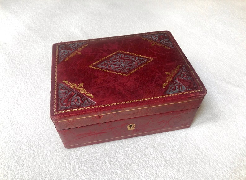 May include: A small, red leather box with gold accents and a decorative design. The box has a keyhole and a hinged lid.