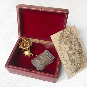 May include: A red velvet lined box with a gold flower brooch and a silver pocket notebook with a floral design. The box is open and the notebook is partially closed. The notebook has the text "Carcassonne La Cite" on the front.