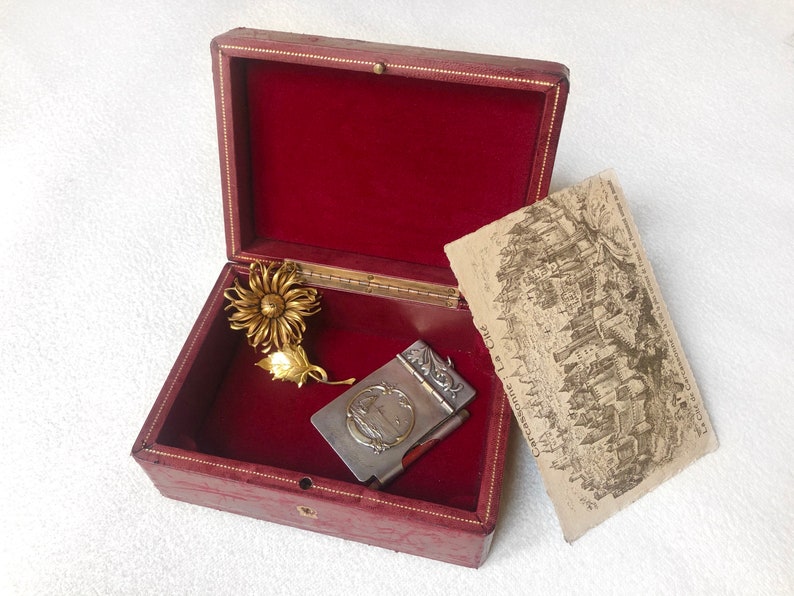 May include: A red velvet lined box with a gold flower brooch and a silver pocket notebook with a floral design. The box is open and the notebook is partially closed. The notebook has the text "Carcassonne La Cite" on the front.