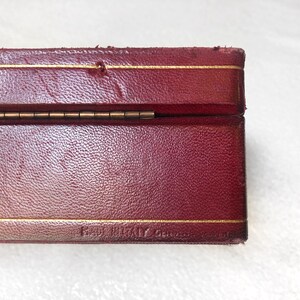 May include: A red leather box with a gold trim and a brass hinge. The box is closed and the bottom of the box is visible. The text "Hand Made in Great Britain" is visible on the bottom of the box.