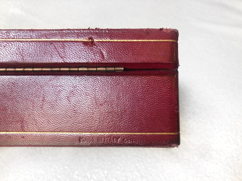 May include: A red leather box with a gold trim and a brass hinge. The box is closed and the bottom of the box is visible. The text "Hand Made in Great Britain" is visible on the bottom of the box.