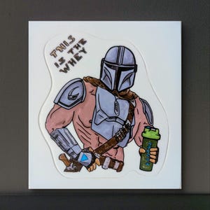 Può includere: Un adesivo con un'illustrazione a cartone animato di un personaggio muscoloso di Mandalorian che tiene una bottiglia di frullato proteico verde. Il testo "This is the whey" è scritto in un font stilizzato sopra il personaggio.