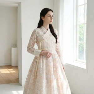 Pode incluir: Um vestido de manga comprida cor pêssego claro com uma sobreposição de renda floral. O vestido apresenta uma gola mandarim, uma fita amarelo pálido e punhos brancos. O vestido tem uma saia ampla e é usado por uma pessoa.