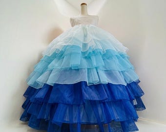 Pode incluir: Vestido degradê em tons de azul, exposto em um manequim. O vestido apresenta uma saia com babados em camadas que vai do azul claro ao azul escuro. A parte superior é branca. Um exemplo marcante de design em camadas.