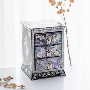 Pode incluir: Um porta-joias preto com três gavetas, apresentando incrustações de madrepérola iridescente com motivos florais e de borboletas. Cada gaveta possui um puxador prateado em forma de borboleta. O exterior é decorado com padrões de conchas.