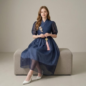 May include: A navy blue dress with white polka dots, featuring sheer short sleeves and a flowing skirt. The dress has a V-neckline and is adorned with a floral brooch and a pink tassel pendant. The model is wearing white heels.