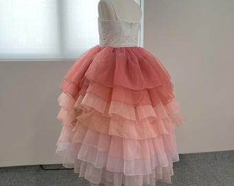 Pode incluir: Um vestido formal em um manequim. O corpete é branco, com alças finas. A saia é em camadas, com um gradiente de cores do rosa ao pêssego ao branco. O vestido foi projetado para uma ocasião especial.