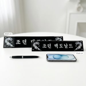 May include: Two black nameplates with white text and dragon designs. The larger plate measures 60x8 cm, and the smaller one is 55x8 cm. A black pen and a smartphone are also on the white surface.