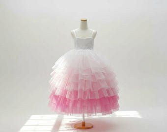 Pode incluir: Vestido de tule branco e rosa ombré em um manequim. O vestido apresenta uma saia em camadas que vai do branco a um rosa vibrante. O corpete tem alças finas. O vestido é exibido em um suporte de madeira sobre um fundo branco.