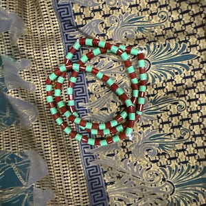 May include: A necklace made of alternating red and light green cylindrical beads. The beads are strung together in a circular design. The necklace is shown on patterned fabric with gold, blue, and white colours.