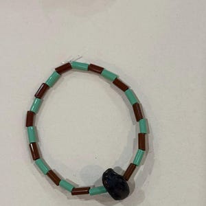 May include: A beaded bracelet featuring alternating brown and turquoise tube beads. A dark blue, irregularly shaped bead serves as a focal point. Additional strands of similar beads are visible in the background.
