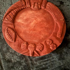 Puede incluir: Un plato decorativo circular de color terracota que presenta un relieve tallado de un rostro humano en la parte superior, una cabeza de elefante en la parte inferior y símbolos tallados adicionales que incluyen una forma de hoja y un patrón circular.