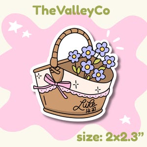 May include: A brown woven basket sticker featuring a bouquet of light purple flowers with yellow centers. The basket is decorated with a light pink ribbon bow and lace trim. The text Luke 12:27 is written on the front. The sticker measures 2x2.3 inches.