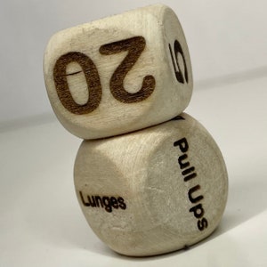 May include: Two wooden dice with black text. The top die has the number 20 on it. The bottom die has the words "Lunges" and "Pull Ups" on it.