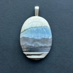 May include: An oval-shaped pendant featuring horizontal bands of color including cream, light blue, gray, and dark brown. The surface has a smooth, polished finish and is topped with a silver-toned metal bail for attachment to a chain or cord.