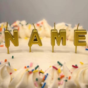 May include: Four gold-colored letter-shaped candles spelling the word NAME, each with a thin metal wick, standing upright on a white frosted cake decorated with a border of piped frosting swirls and colorful rainbow sprinkles.