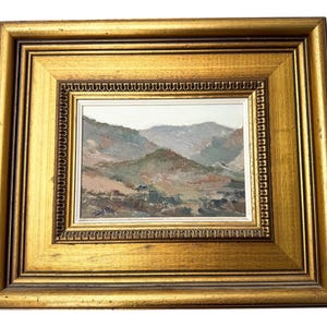 May include: A framed landscape painting with a gold-colored ornate frame. The painting depicts a mountain range in muted tones of green, brown, and gray. The frame has a double border with a detailed inner edge.