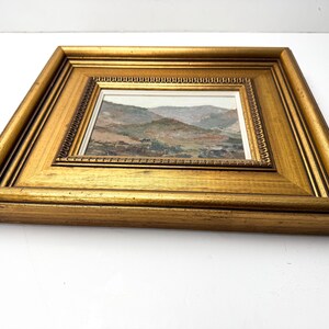 May include: A framed landscape painting with a gold-colored wooden frame. The painting depicts a mountainous scene with muted colors of green, brown, and gray. The frame has a detailed, ornate design.