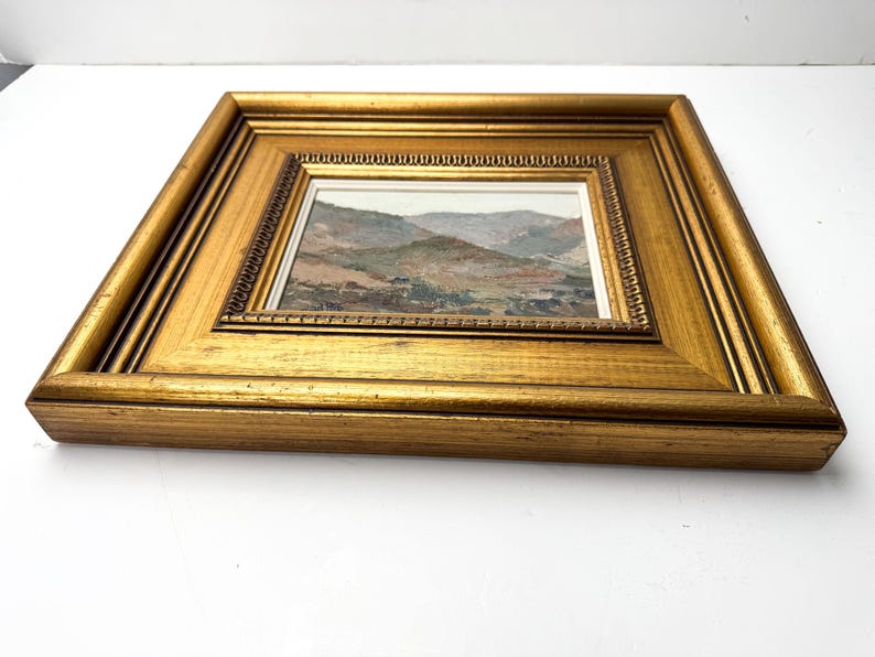 May include: A framed landscape painting with a gold-colored wooden frame. The painting depicts a mountainous scene with muted colors of green, brown, and gray. The frame has a detailed, ornate design.