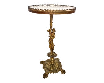 May include: An antique-style side table in a gold finish. The table has a round top with a decorative edge. A sculpted figure supports the tabletop. The base has a detailed, floral design. The table is a decorative accent piece.