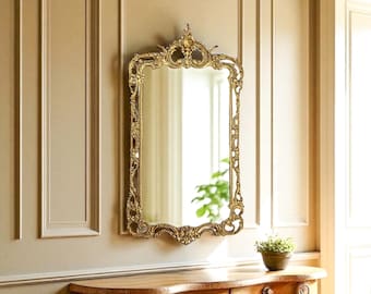 May include: A rectangular wall mirror featuring an ornate gold-colored frame with intricate scrollwork and floral carvings. The mirror has a decorative crest at the top and a symmetrical design along the sides and bottom edges.