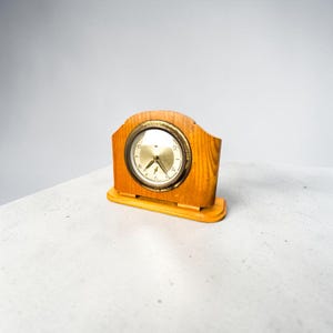 Op de afbeelding: Een vintage houten klok met een lichtbruine afwerking. De wijzerplaat is goudkleurig met zwarte cijfers en wijzers. De klok heeft een rechthoekige basis en een gebogen bovenkant. Afmetingen: ongeveer 15 cm breed en 10 cm hoog.