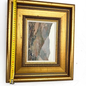 May include: A framed landscape painting with a gold-colored wooden frame. The painting features a mountain scene with muted tones of brown, gray, and white. The frame measures approximately 17 inches in length.
