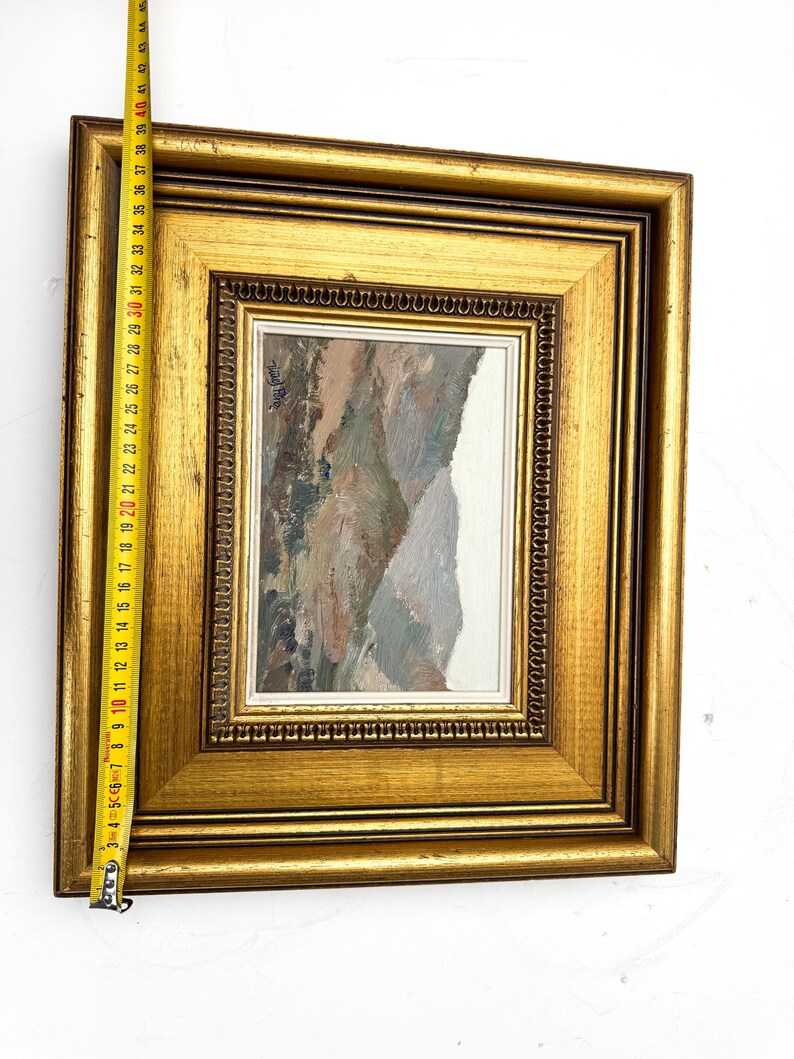 May include: A framed landscape painting with a gold-colored wooden frame. The painting features a mountain scene with muted tones of brown, gray, and white. The frame measures approximately 17 inches in length.