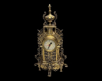 May include: An ornate gold-toned metal mantel clock featuring a circular white face with black Arabic numerals and black hands. The clock is encased in a highly detailed, intricate frame with scrollwork, vertical bars, and a decorative finial top.