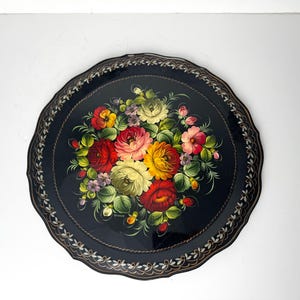 Könnte beinhalten: Runder, schwarzer dekorativer Tablett mit gewelltem Rand und einem Blumengesteck-Design. Das Bouquet zeigt rote, gelbe, rosa und violette Blumen mit grünen Blättern. Der Rand hat eine weiße und goldene Verzierung.