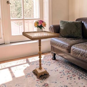 Pode incluir: Uma mesa lateral decorativa com um tampo quadrado e uma borda dourada ornamentada. A mesa tem uma coluna esguia e esculpida e uma base quadrada. Um vaso de flores coloridas está sobre a mesa, ao lado de um sofá de couro marrom.