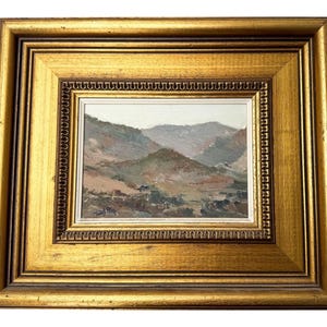 May include: A framed landscape painting featuring a mountain scene in muted tones of brown, green, and gray. The painting is set within a gold-colored, ornate frame, adding a touch of elegance to the artwork.