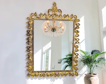 May include: A rectangular wall mirror featuring an ornate gold frame with intricate scrollwork and a central decorative crest at the top. The mirror reflects a multi-armed chandelier and a portion of a room with a potted green plant nearby.