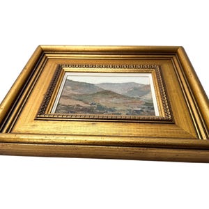 May include: A framed landscape painting with a gold-colored wooden frame. The artwork depicts a mountainous scene with muted earth tones. The frame has a detailed, ornate design.