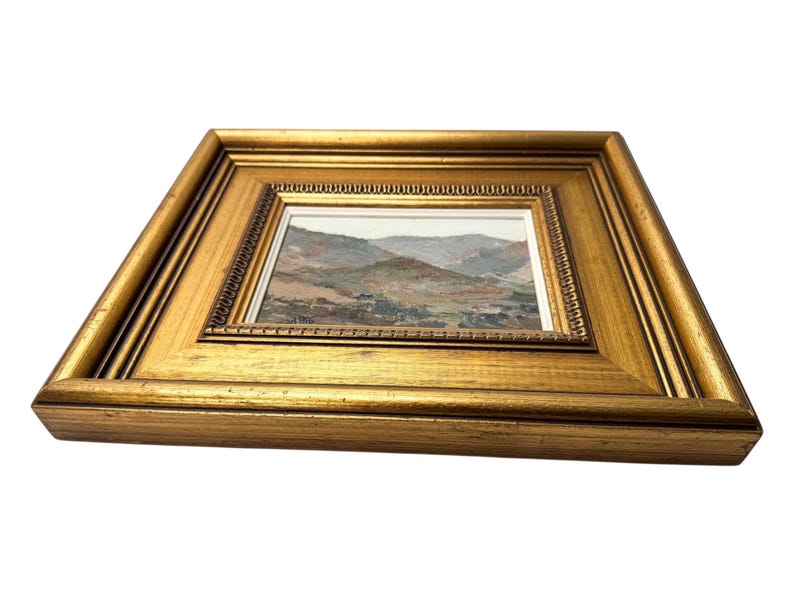 May include: A framed landscape painting with a gold-colored wooden frame. The artwork depicts a mountainous scene with muted earth tones. The frame has a detailed, ornate design.