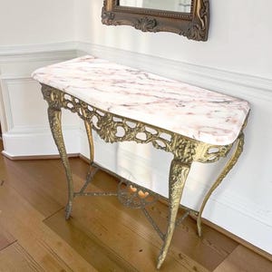 May include: A console table featuring a rectangular pink and white marble top supported by an ornate, curved brass metal base with intricate floral and scrollwork detailing along the apron and four slender, decorative legs.