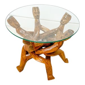 May include: A unique, handcrafted coffee table with a clear glass top supported by a carved wooden base. The base features figures with carved faces and outstretched arms, creating an artistic and functional piece.