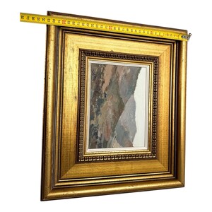 May include: A framed landscape painting with a gold-colored ornate frame. The painting depicts a mountain scene with muted tones of brown, gray, and white. A measuring tape shows the frame is approximately 15 inches wide.
