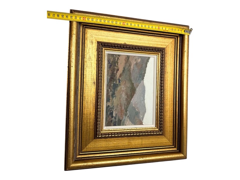 May include: A framed landscape painting with a gold-colored ornate frame. The painting depicts a mountain scene with muted tones of brown, gray, and white. A measuring tape shows the frame is approximately 15 inches wide.