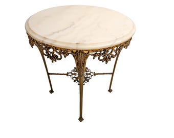 May include: A small, round side table with a white marble top. The table has an ornate, gold-coloured metal frame with floral and scrollwork details. The table's legs are slender and slightly curved.