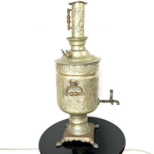 May include: An antique silver samovar with intricate floral designs. The samovar has a spout and a handle on the side, and a tall, ornate top. It is sitting on a black base.