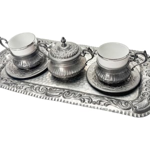Puede incluir: Un juego de té plateado sobre una bandeja decorativa. El juego incluye dos tazas de té con platillos y un azucarero con tapa. La bandeja y el juego tienen detalles ornamentados.