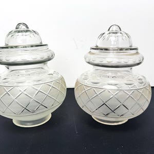 Può includere: Due lampadari in vetro trasparente con un motivo a rombi nella metà inferiore e una parte superiore arrotondata. I lampadari hanno un design decorativo e un piccolo terminale arrotondato sulla parte superiore. Sono su una superficie scura.