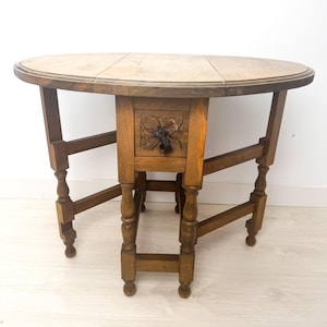 Op de afbeelding: Een bruine ovale klaptafel van hout met een gegraveerde lade en gedraaide poten. De tafel heeft een eenvoudig ontwerp en is perfect voor een kleine ruimte.