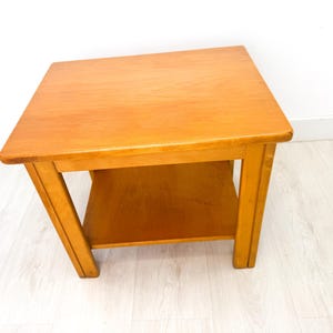 Peut inclure: Une table d'appoint carrée en bois avec une finition brun clair. La table a une étagère inférieure et quatre pieds robustes. La table mesure environ 51 cm de large, 51 cm de profondeur et 51 cm de haut. La table est en bois.