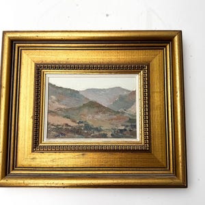 May include: A framed landscape painting featuring a mountain range in muted tones of brown, green, and gray. The artwork is encased in a gold-colored wooden frame with a detailed inner border, creating a classic aesthetic. The painting is a small size.